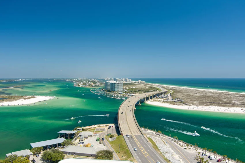 A bridge crosses an emerald-green inlet in Orange Beach, Alabama, with passing boats, white-sand shores, and waterfront buildings visible from above.