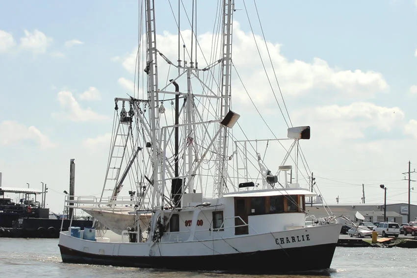 A shrimp boat named Charlie travels through the harbor in Venice, Louisiana, with tall rigging above its white cabin and dark hull.