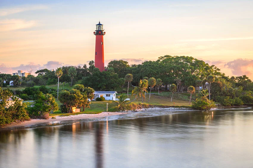 Aerial view of Jupiter, Florida, USA with the iconic inlet lighthouse illuminated at dawn, soft light reflecting on the calm water.