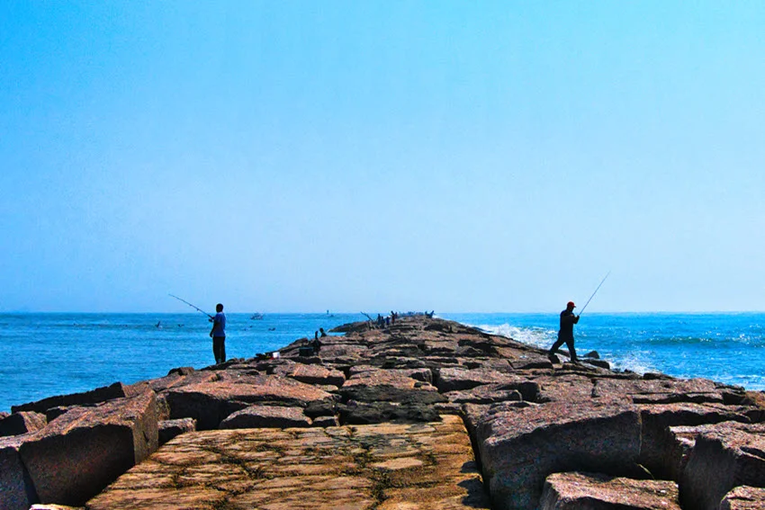Anglers standing on a long rocky jetty casting lines into the calm blue water under a clear sky.