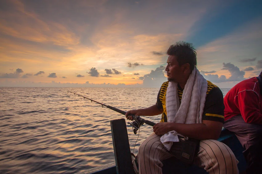 Angler on a fishing boat at sunrise, watching the spread and scanning the horizon over calm offshore water.