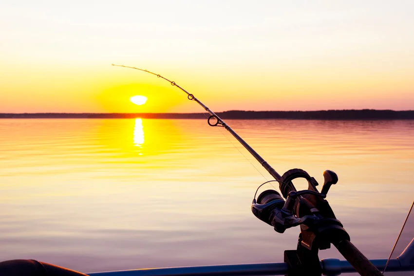 Close-up shot of a spinning reel in a rod holder at sunset, set up for trolling or drift fishing in open water.