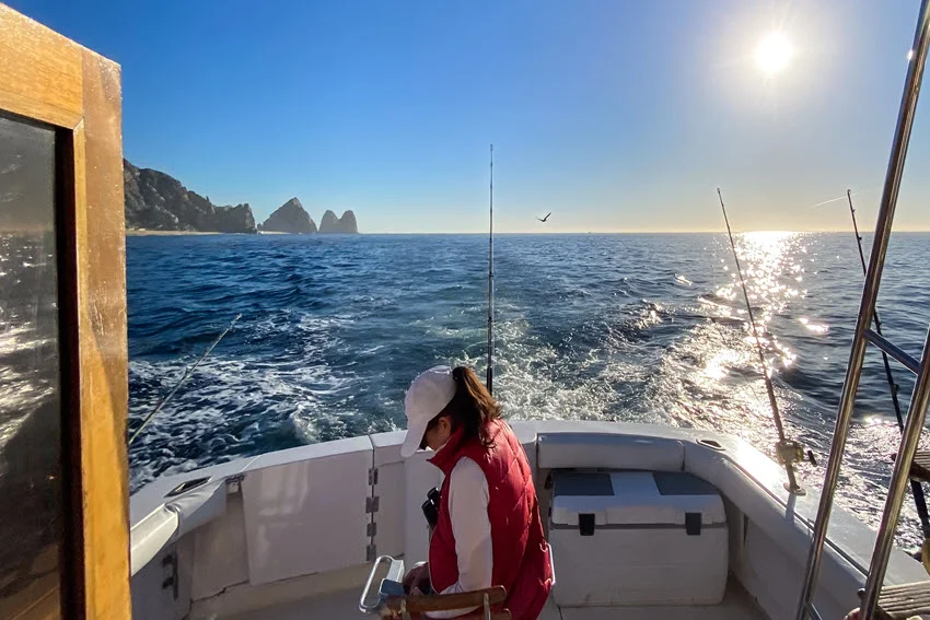 Woman on a charter boat fighting a fish on heavy tackle, braced at the rail with offshore water and wake behind the boat.