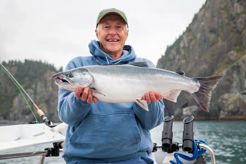 Angler holding a Salmon in Alaska with rugged mountains behind, a destination style scene tied to remote runs and cold water fisheries.