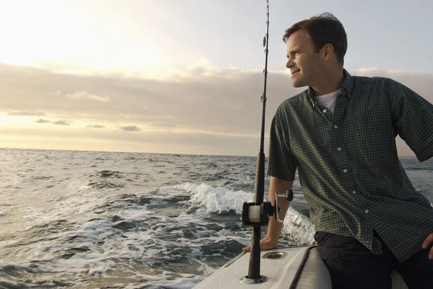 An angler leaning on the edge of the boat, standing next to a fishing rod and gazing into the distance toward the golden sunset horizon.