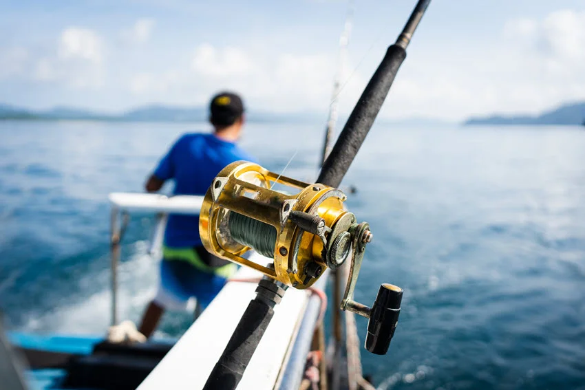 Focused shot of golden fishing reels attached to a moving fishing boat, with an angler and the blue sea in the background.
