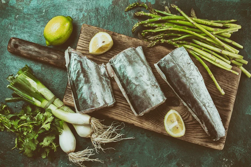 Top view of raw Sawfish fillets, placed on wooden board with asparagus, lemon and herbs.
