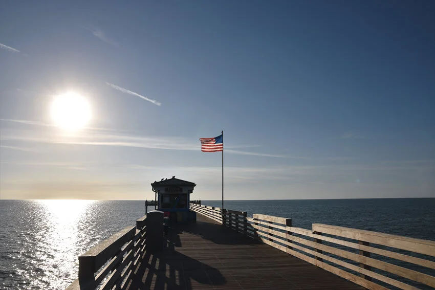 Scenic view of a Florida Gulf Coast fishing pier at sunrise with an American flag, calm water, and long shadows across the wooden deck.