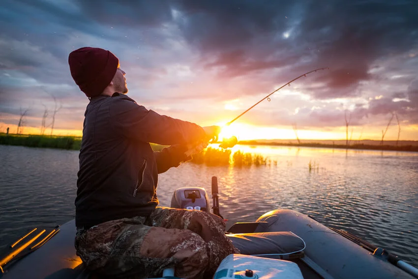 Wide shot of an angler fishing from an inflatable boat in the bay, silhouetted against the warm colors of the sunset.