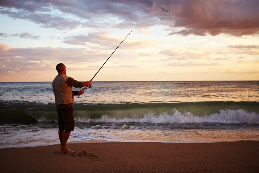 A back view of an angler performing flipping and pitching from an ocean shore in the early morning, with a scenic sky above that transitions from deep blue to purple near the horizon.