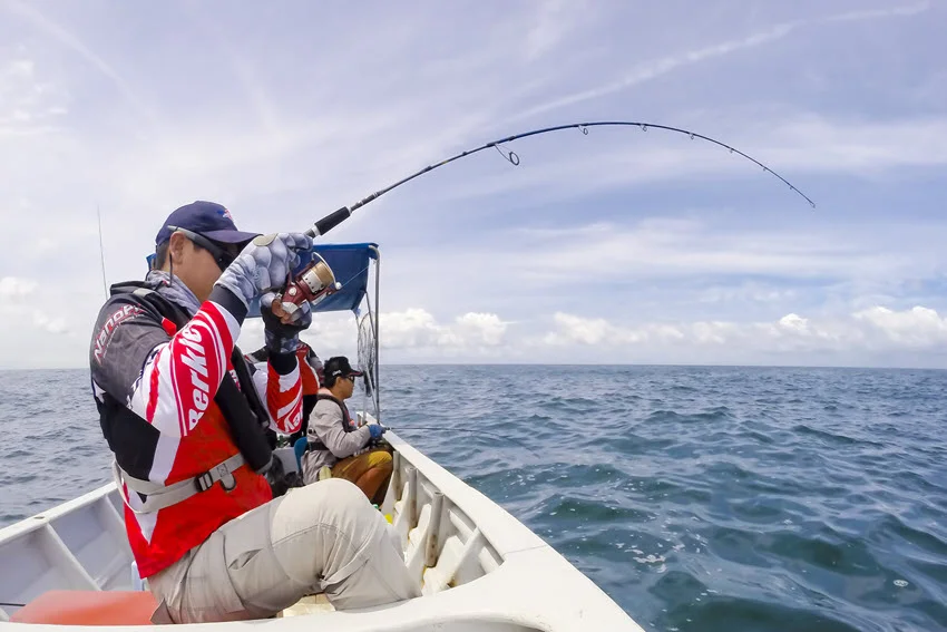 A side view of an angler seated on a small boat, performing jigging fishing offshore, with open water stretching out to the horizon.