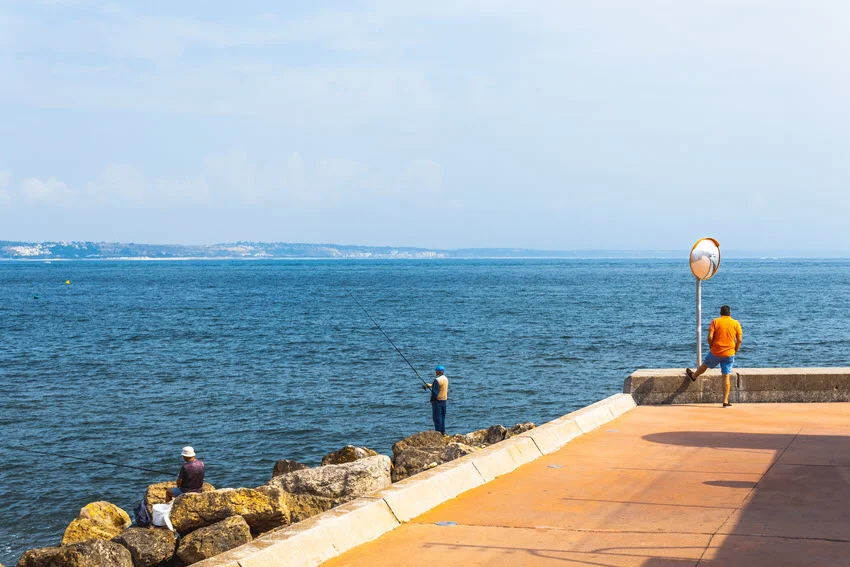 Wide shot of two anglers standing on a rocky shore, performing sight fishing under a clear, sunny sky.