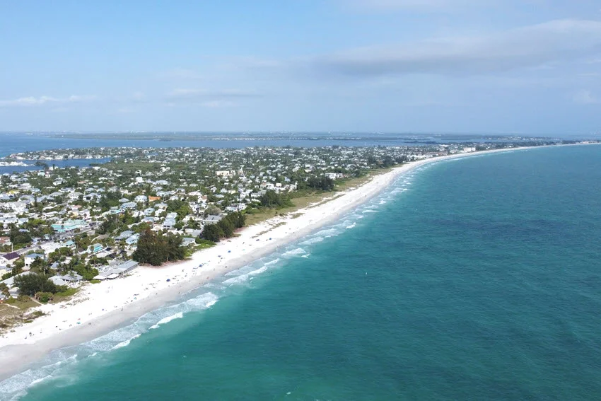 An aerial view of Anna Maria Island on Florida&rsquo;s Gulf Coast, showing its white sandy beaches, turquoise shoreline, and coastal neighborhoods under a clear blue sky.