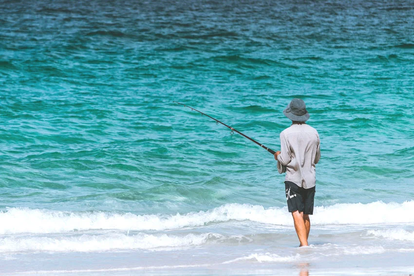Angler casting from the beach into clear surf, working the shoreline trough and wash where bait moves close and predators often cruise in shallow water.