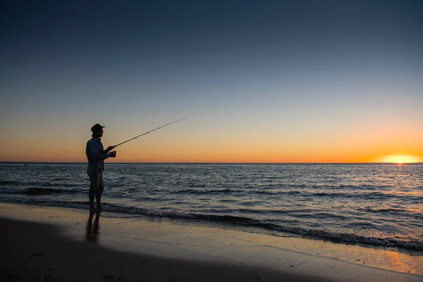 Angler silhouette casting from the beach at sunset, a classic low-light window when predators often move shallow along the surf line and nearshore breaks.