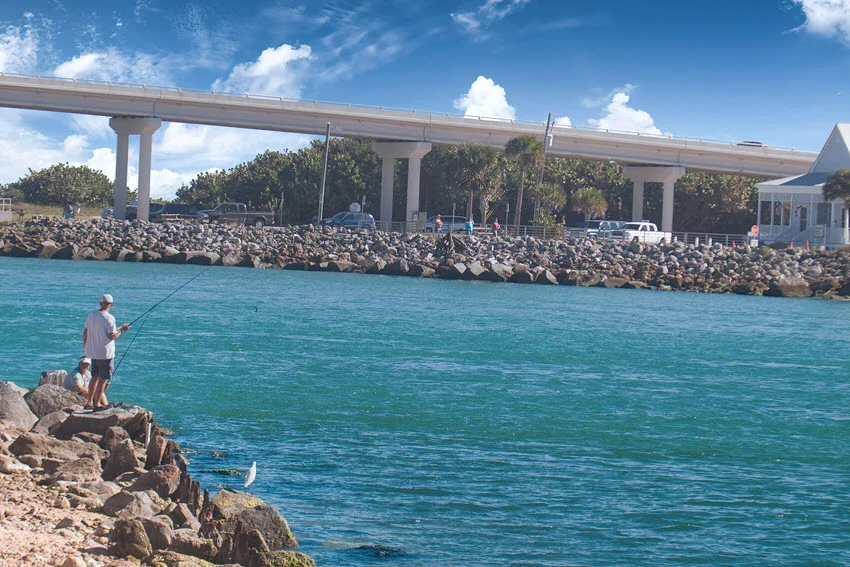 Shoreline view of an angler casting near rocky jetty structure at Sebastian Inlet, Florida, on a bright day.