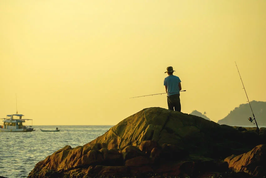 Fly-fishing angler casting a long line at sunset from the shoreline, with gentle surf and golden light across the water.