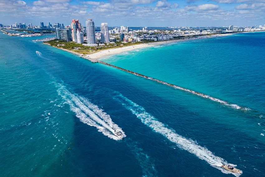 Aerial shot of Miami's coastline and Biscayne Bay, with boats leaving white wakes through bright turquoise water.