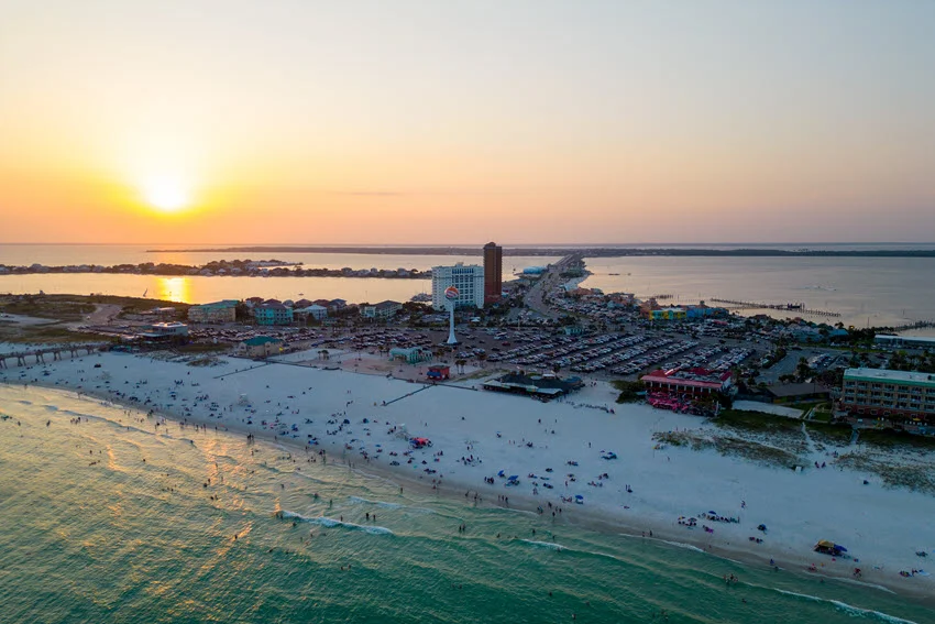 Aerial view of Pensacola Beach, Florida at sunset, with beach crowds, warm sky colors, and the shoreline fading into the distance.