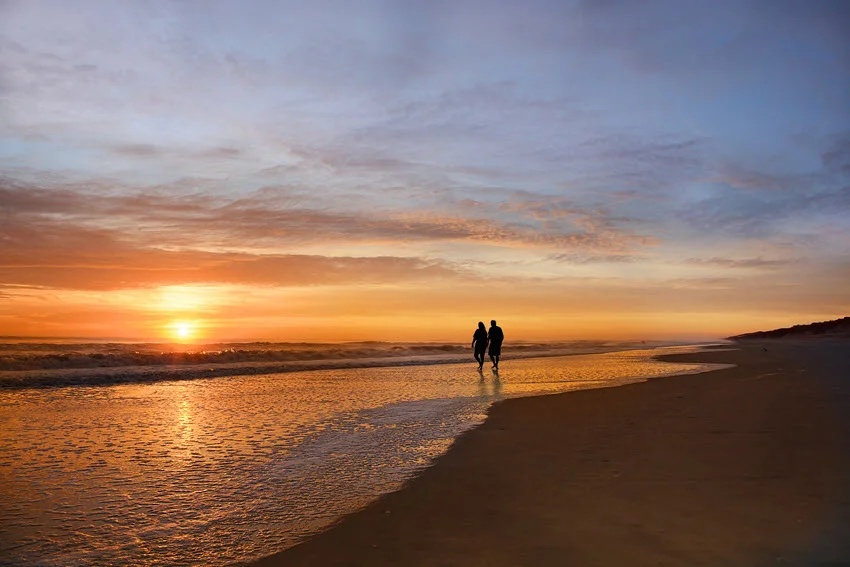 Senior couple walking along a North Carolina beach at sunset, with wet sand reflecting the sky and gentle waves nearby.