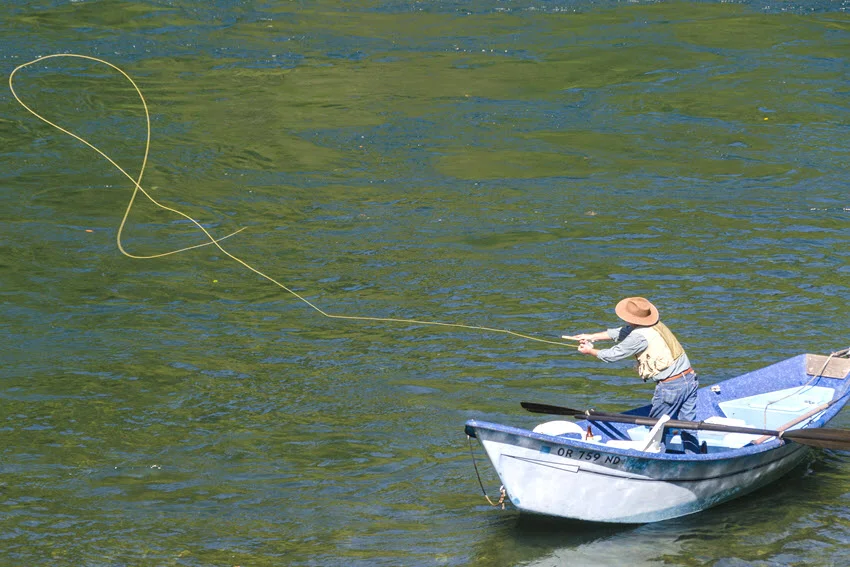 Angler fly casting from a drift boat, with a long loop of fly line suspended above the rippled green water.