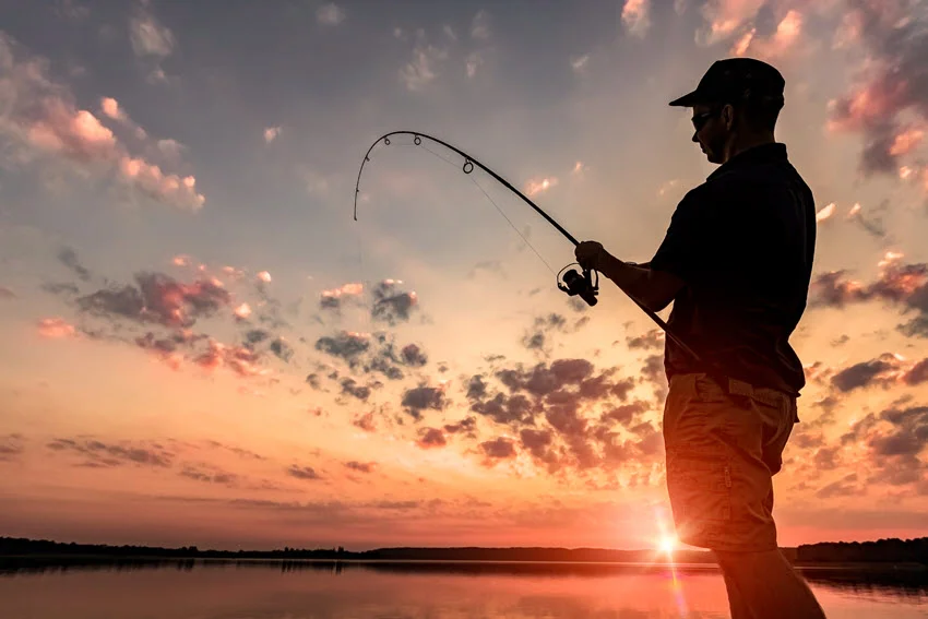 Silhouetted angler holding a bent spinning rod beside a lake, with the sun low on the horizon beneath orange clouds.