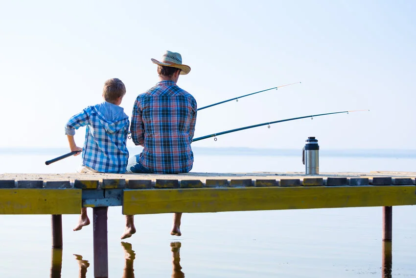 An adult angler and a child sit side by side on a wooden pier, holding fishing rods above calm water.