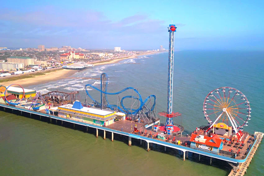 Aerial view of Galveston's Pleasure Pier, with a Ferris wheel and roller coaster extending over Gulf waters beside the sandy beachfront.
