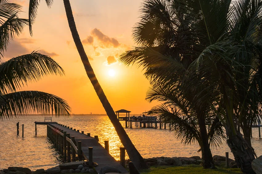 Golden sunset over Florida's Indian River, with wooden docks extending into the water and silhouetted palms framing the shoreline.