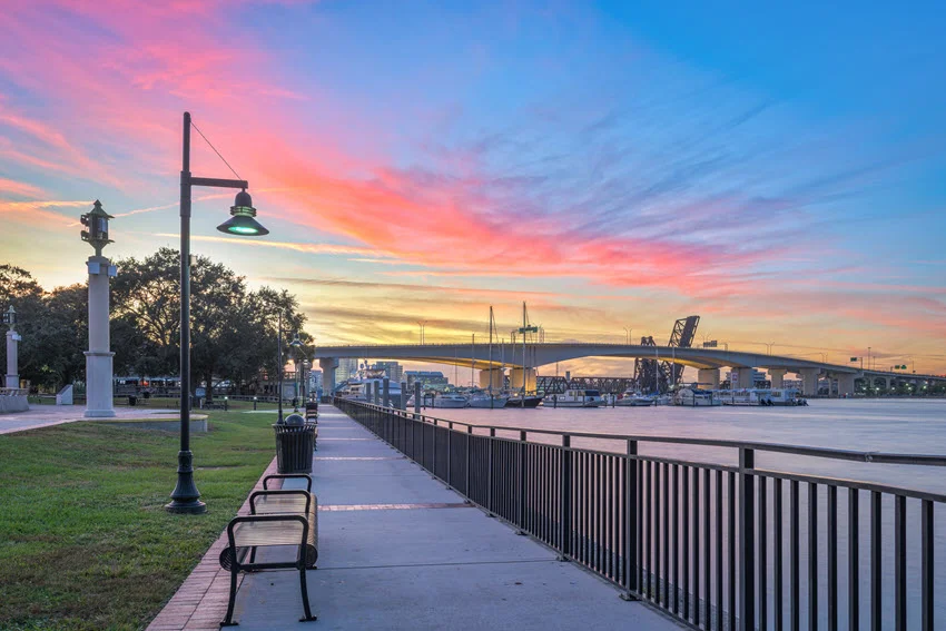 Benches and lamps line Jacksonville's Riverwalk beside the St. Johns River, with bridges beneath a pink and blue dusk sky.