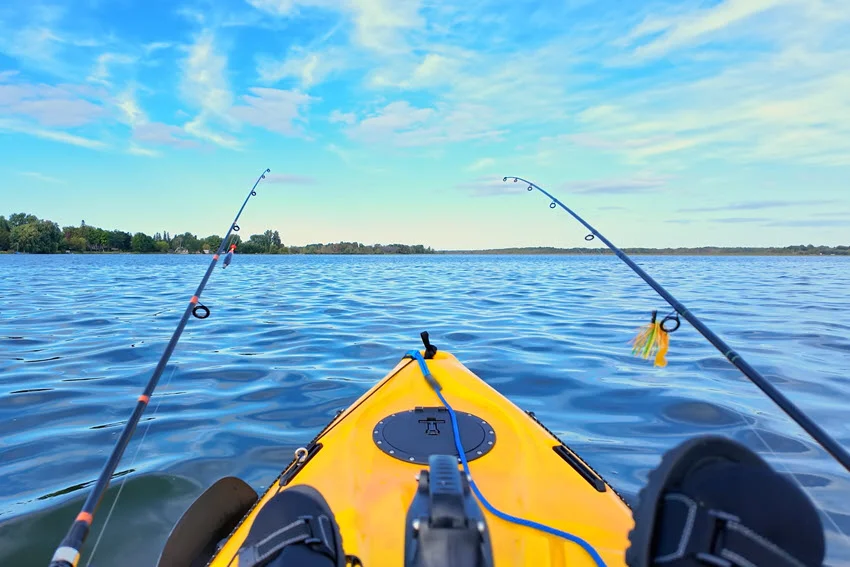 Two fishing rods flank the bow of a yellow pedal kayak on a lake, with a tree-lined shoreline in the distance.