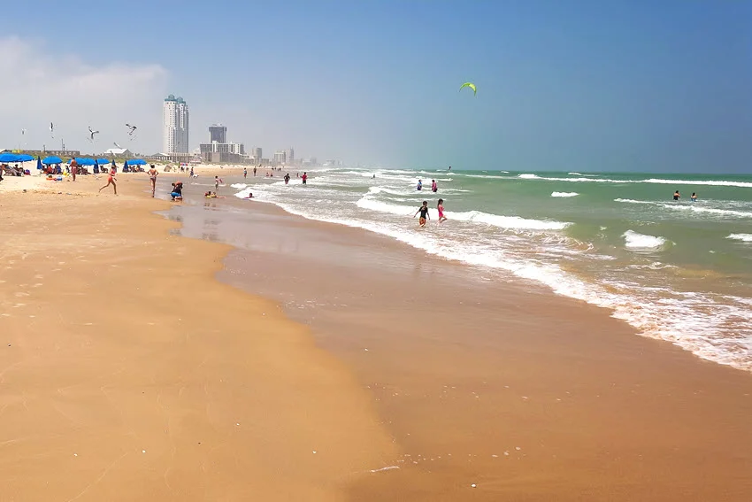 Beachgoers walk and swim along South Padre Island, Texas, as waves wash onto the sand beside beach umbrellas and distant hotels.