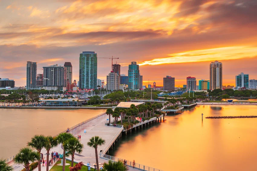 Downtown St. Petersburg, Florida, rises beyond a palm-lined waterfront walkway, with orange clouds and warm reflections spreading across the water.