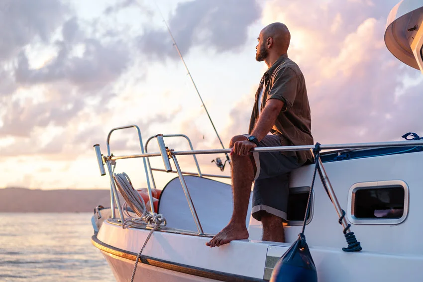 Angler standing with fishing rod on a side of its white sailboat, fishing in open sea, and gazing into sunset.