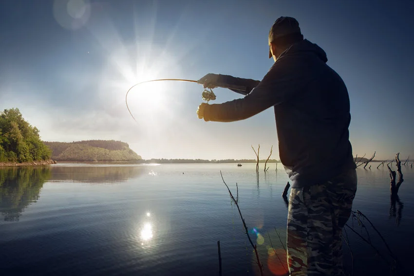 Angler stands on a lake shore, casting his rod into calm water during sunrise.