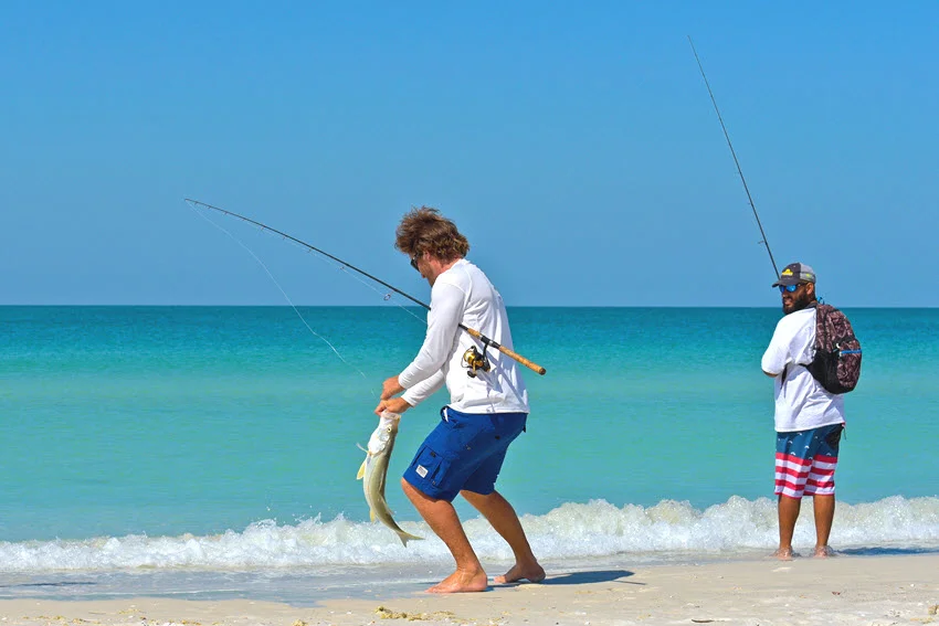 Back view of two anglers standing on a sandy beach, casting lines into light blue water.