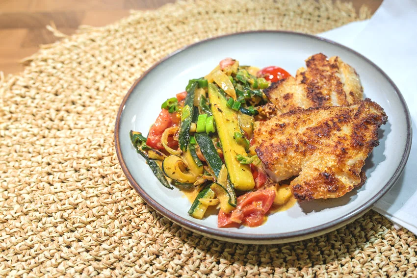 Crispy fried Redfish fillet served with zucchini, tomatoes, and leek on a gray plate.