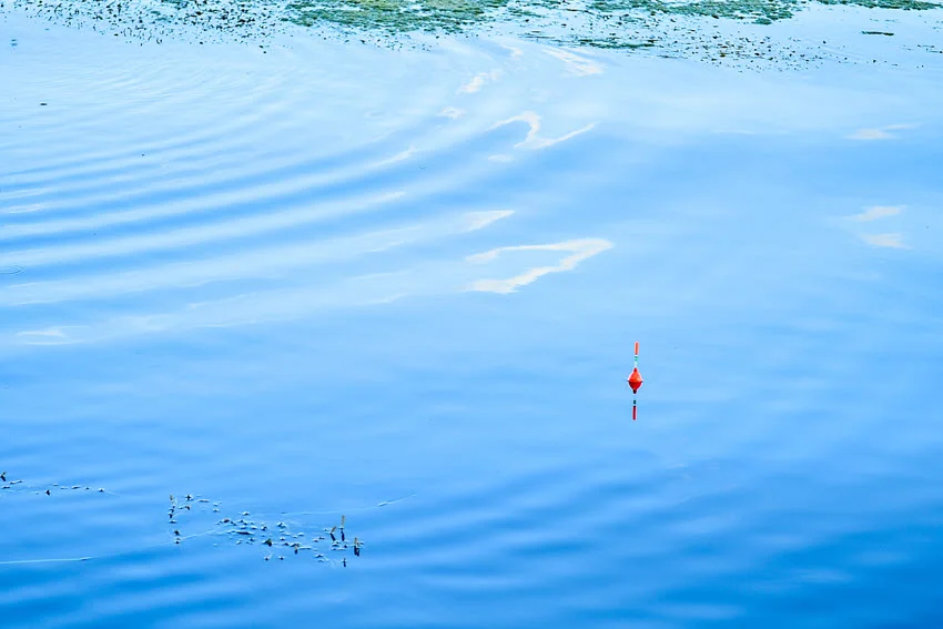 Distant shot of an angler's red float from a fishing rod drifting on blue water reflecting the sky.