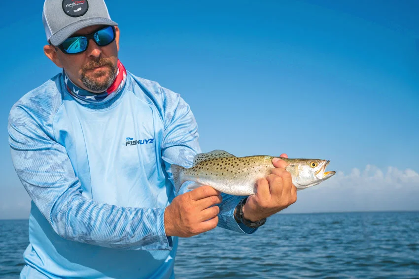 View of an angler holding a Spotted Seatrout over open water, showing the fish spots, tail, and body shape in clear daylight.