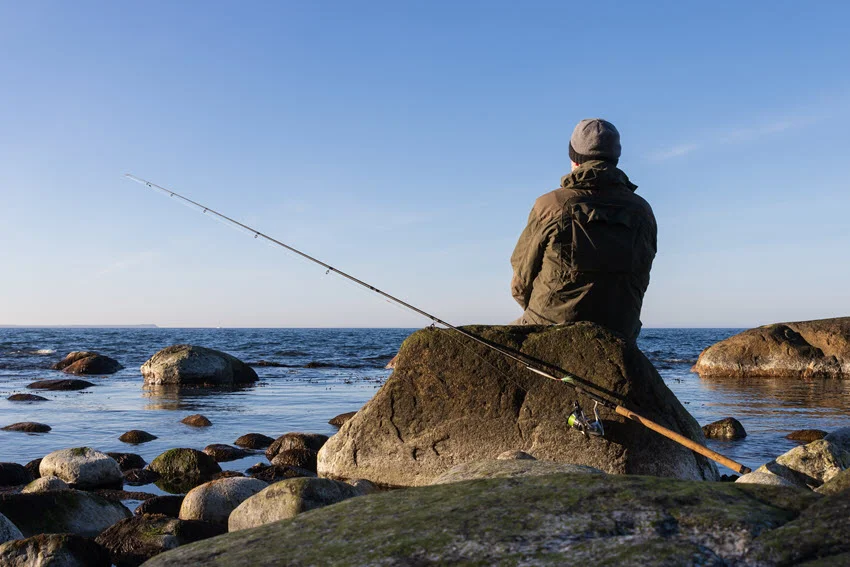 Wide shot of an angler fishing from a rocky shoreline with a long rod pointed over the water during calm, clear conditions.