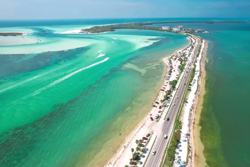 Aerial view of turquoise coastal water, sandbar colors, and a long causeway along a tropical shoreline on a bright, clear day.