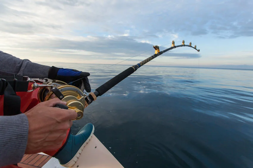 A close shot of an angler's hand firmly holding a fly fishing rod on a boat, with subtle water reflections in the background.