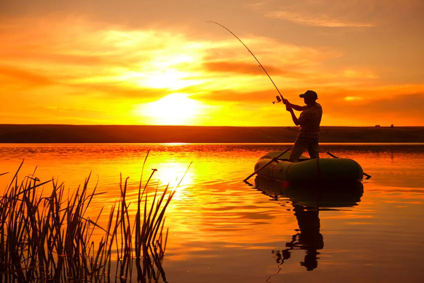 Silhouette of a man standing in a boat, fishing at sunset, with a vibrant orange sky reflecting on the calm water surface, and reeds in the foreground.