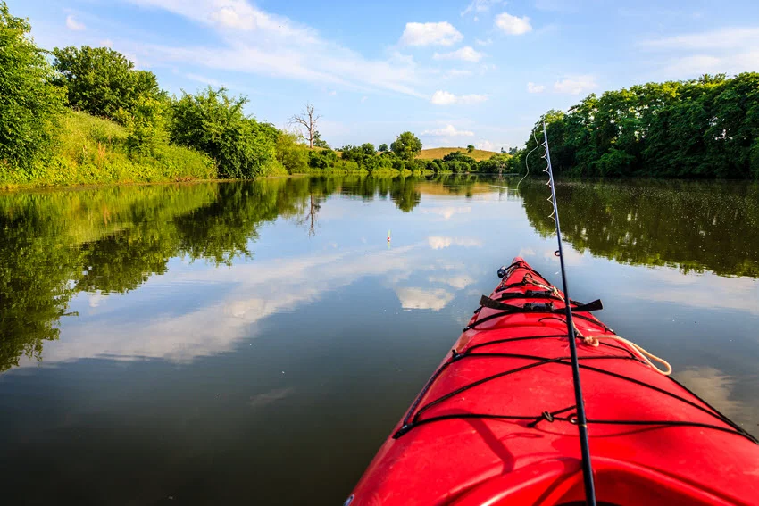 View from a red kayak on calm waters, line cast for fishing, surrounded by lush greenery and a clear sky reflected on the water's surface.
