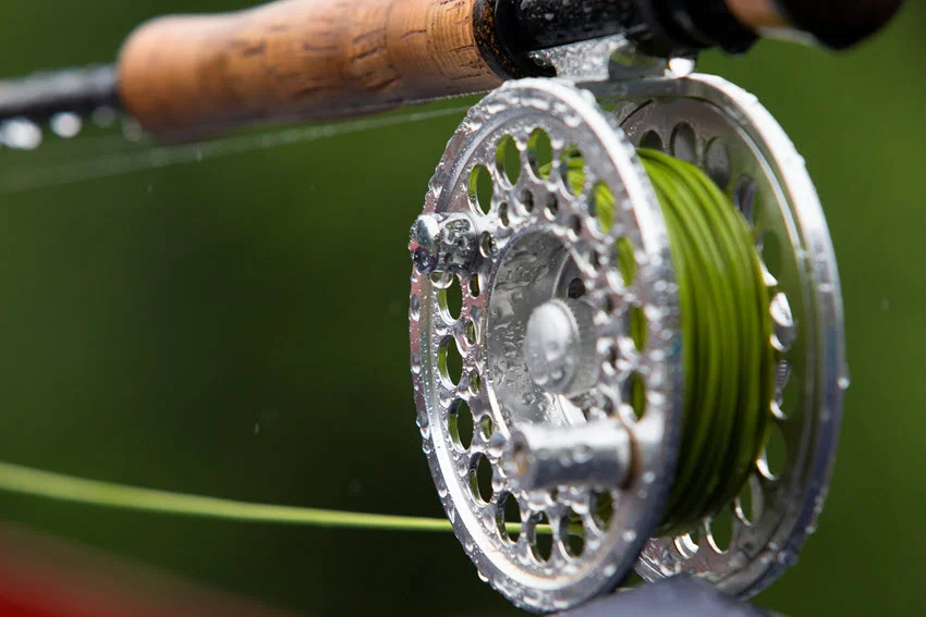 Close-up view of a fly fishing reel with water droplets spinning off the line, with a blue-green background visible.
