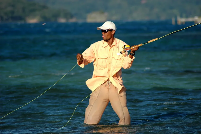 Angler standing in shallow flats water casting a fly rod, dressed in a light shirt and cap, preparing to target Tarpon in clear blue tropical shallows.