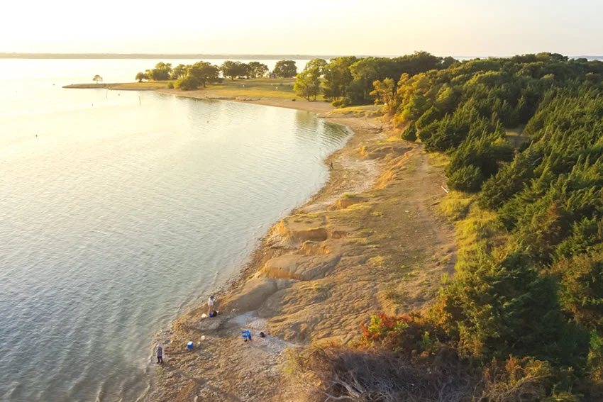 Aerial view of the sandy shoreline at Ticky Creek Park, located at the northern end of Lake Lavon in Princeton, Texas, showing people bank fishing along the water's edge.