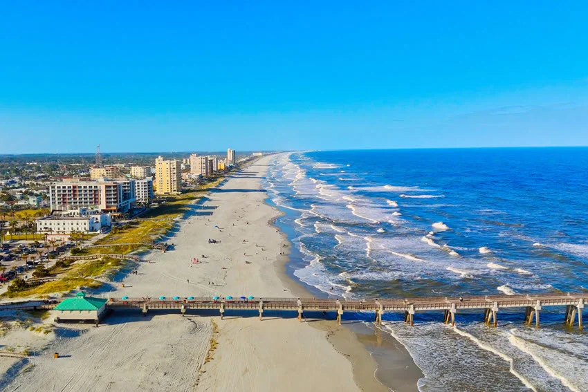 Aerial view of Jacksonville Beach, Florida with a long sandy shoreline, rolling surf, and beachfront buildings along the coast.