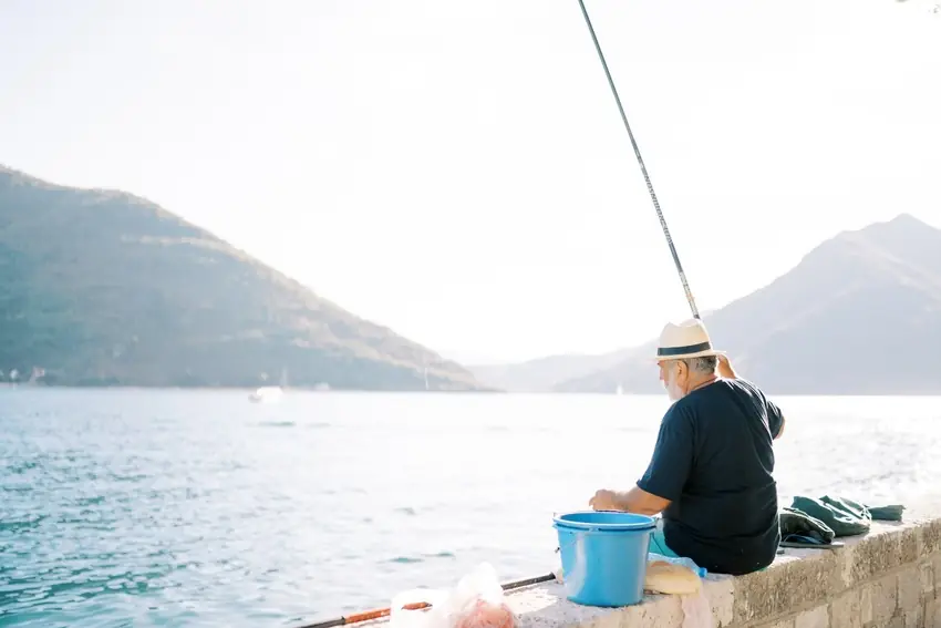 An Angler fishes from a stone seawall overlooking calm coastal water, with steep mountains rising in the background.