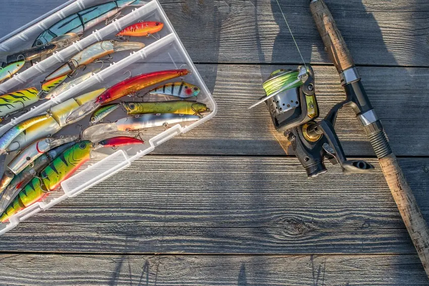 An open tackle box filled with colorful lures sits beside two fishing rods and reels on a weathered wooden surface.